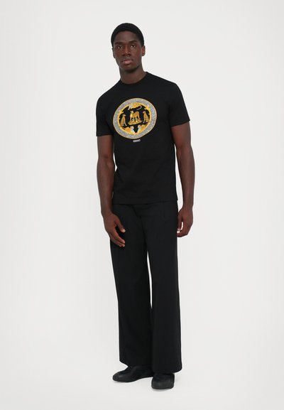 Un tânăr stă în picioare, purtând pantaloni negri, adidași negri și un tricou negru cu un design circular de animal auriu pe piept.