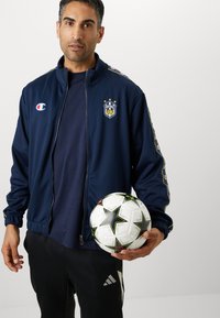 Veste zippée bleu marine avec des emblèmes et des rayures multicolores ; tenant un ballon de football blanc avec un motif d'étoiles noires ; pantalon noir avec logo.