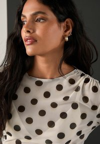 Kvinde med langt mørkt hår iført guldøreringe og en gennemsigtig hvid top med store sorte prikker, der kigger til venstre.