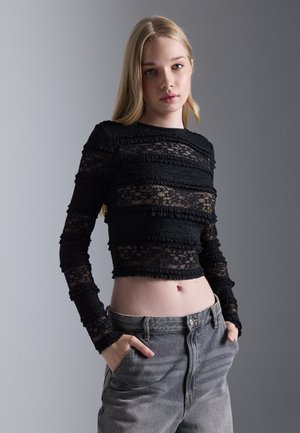 Svart langermet topp med teksturerte blondepaneler, med volanger og en cropped design, kombinert med høyhalsede grå jeans.