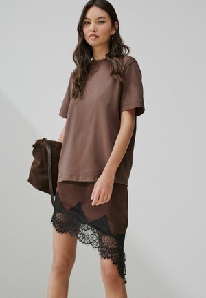 Femme portant un haut marron ample à manches courtes et une jupe asymétrique marron avec un bord en dentelle noire, tenant un sac assorti en suède marron.