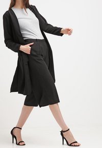 Cardigan largo negro sobre una camiseta a rayas grises y blancas, combinado con culottes negros y sandalias de tacón alto con tiras negras. Textura lisa.