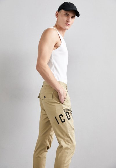 Pantaloni cargo beige con testo stampato nero "ICON" sul lato, indossati con una canotta bianca e un berretto nero, che mostrano un design attillato.