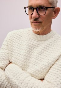 Weißer Strickpullover mit strukturiertem Muster und gerippten Bündchen. Getragen mit quadratischen schwarzen Brillen, vor einem hellen Hintergrund.
