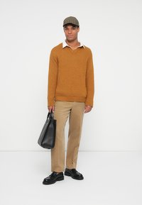 Μουσταρδί πουλόβερ με υφή, λευκή πουκαμίσα, μπεζ παντελόνι, μαύρα δερμάτινα παπούτσια και καπέλο με καρό σχέδιο. Κρατάει μια μαύρη τσάντα tote στο χέρι.