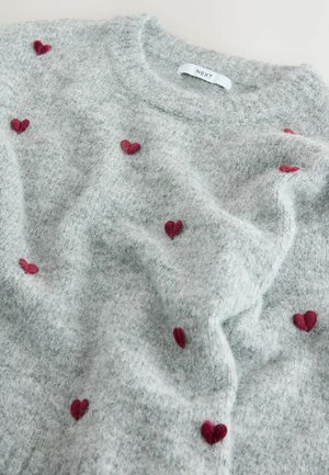 Světle šedý svetr s jemnou texturou, s červenými vyšívanými srdíčky rozptýlenými po látce. Štítek u výstřihu říká "NEXT".