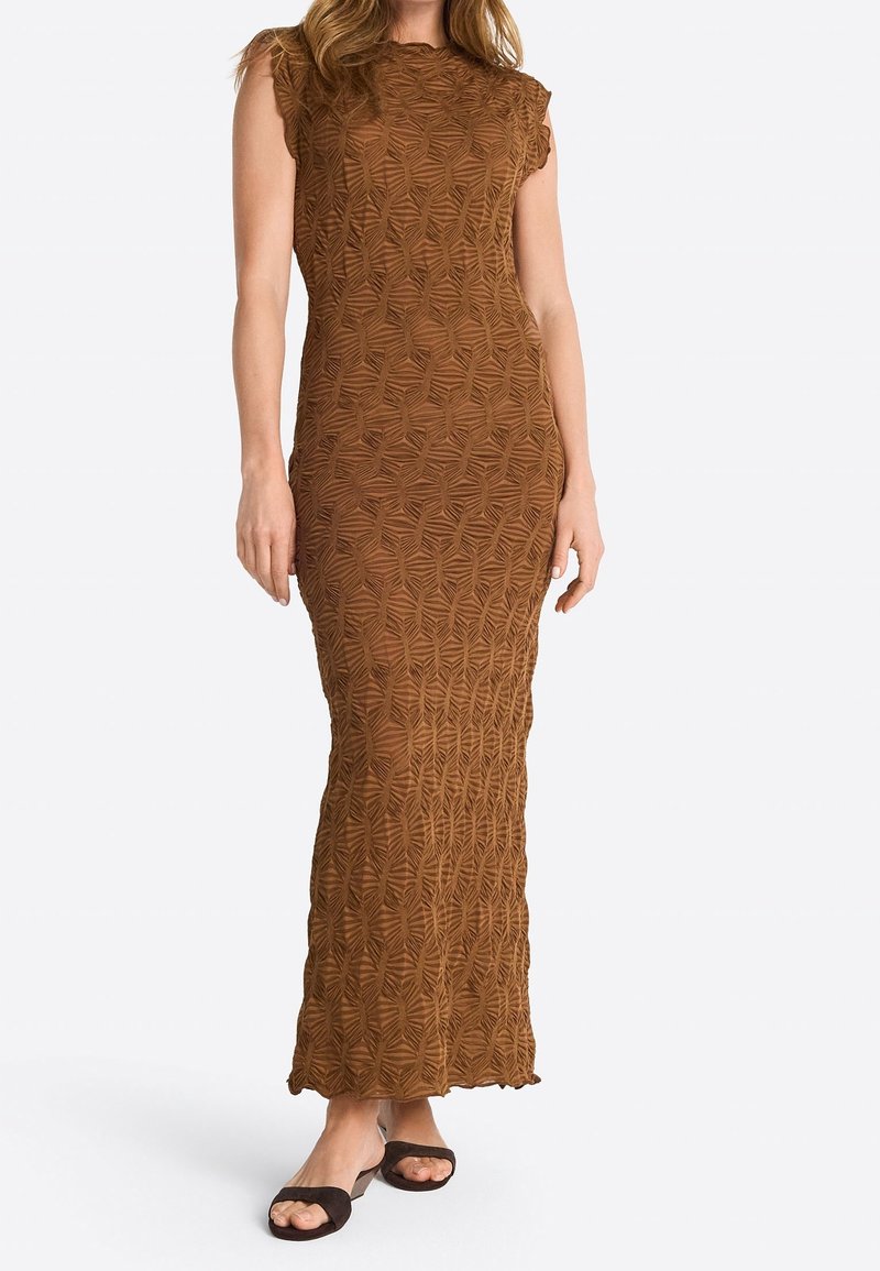 Femme portant une robe maxi marron sans manches texturée avec des motifs géométriques et des sandales noires à enfiler sur un fond uni.