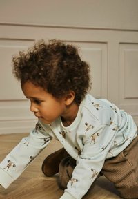 Kährupäise väike laps, kes kannab valget sweatshirt'i, millel on tõstmisnööride trükk ja pruunid püksid, roomab siseruumides puidust põrandal.