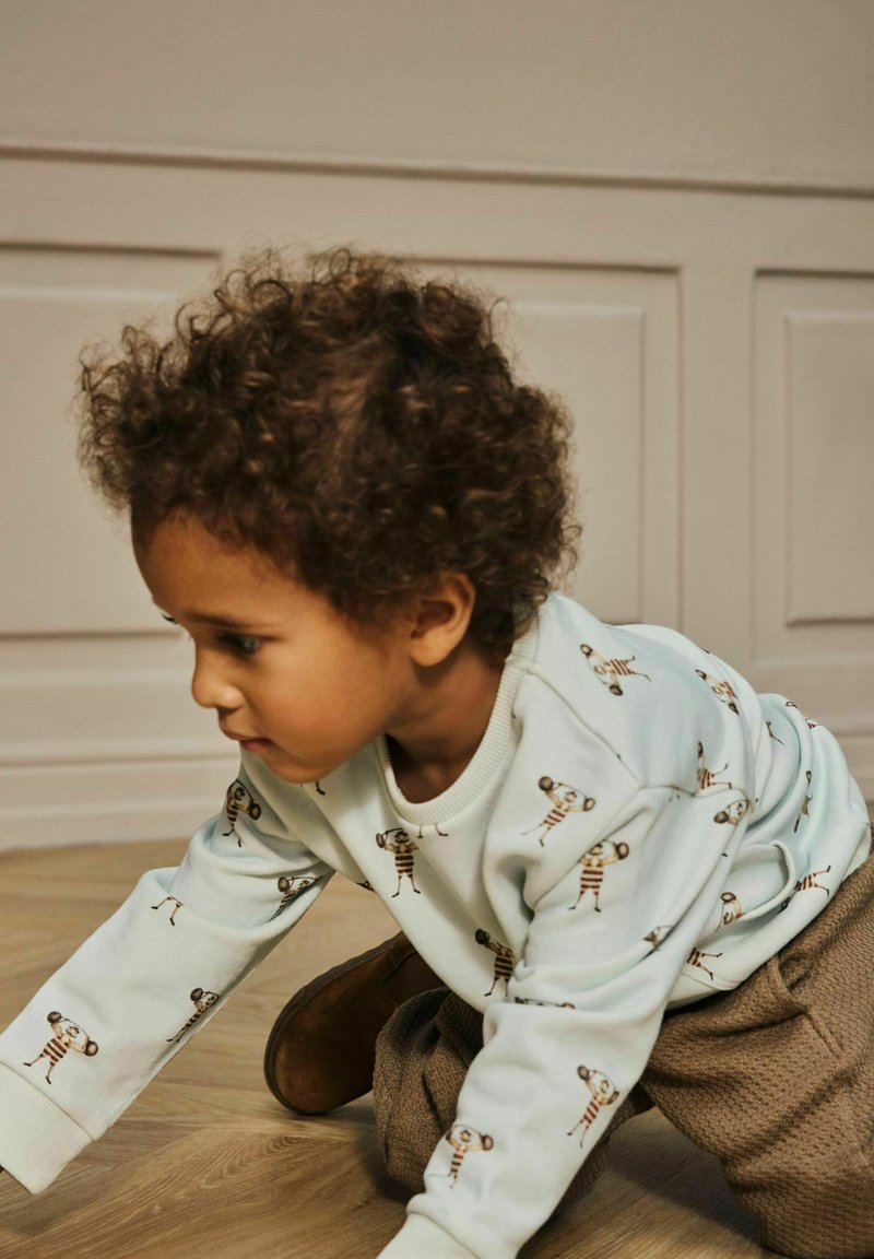 Kährupäise väike laps, kes kannab valget sweatshirt'i, millel on tõstmisnööride trükk ja pruunid püksid, roomab siseruumides puidust põrandal.