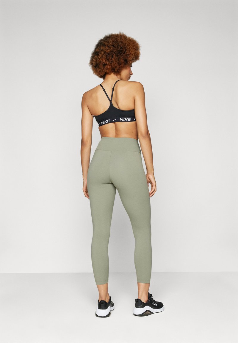 Zwarte sportbeha met wit logo, gecombineerd met olijfgroene leggings. Model draagt zwarte sportschoenen. Focus op pasvorm en stoftextuur.