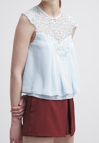 Vaaleansininen hihaton toppi, jossa on monimutkainen pitsiysitys kaula-aukolla ja löysä, virtaava muotoilu. Yhdistetty burgundinpunaisiin shortseihin.