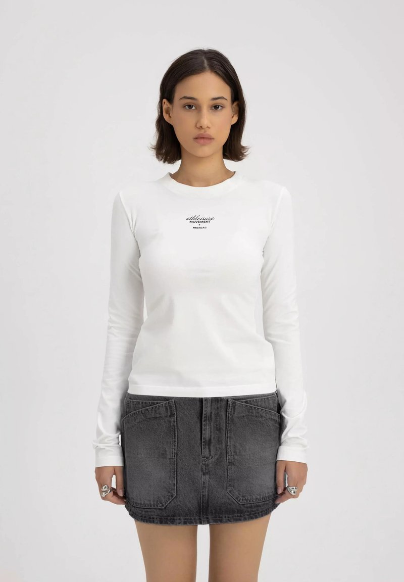 Weißes Langarmshirt mit einem schwarzen Logo auf der Brust, kombiniert mit einem kurzen, dunklen Jeansrock mit Gesäßtaschen.