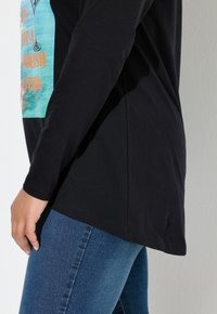 Sort langærmet t-shirt med trykt grafisk design, der forestiller et cocktailglas og tekst. Kombineret med blå denimjeans.