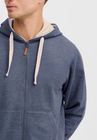 Navy zip-up hoodie made from soft fabric, featuring a beige-lined hood, cream drawstrings, and front pockets. Subtle texture throughout.