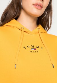 Gelbe Kapuzenjacke mit Kordelzug, ausgestattet mit einem Logo, das "TOMMY JEANS" liest. Glatte Textur und kontrastierende Akzentfarben im Logo.