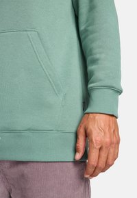 Hoodie verde in cotone con tasca a marsupio, polsini e orlo a costine. Tessuto di trama, cuciture piatte e un piccolo dettaglio con etichetta sono visibili.