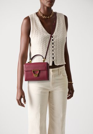 Small burgundy leather handbag with a top handle, gold hardware, and a detachable strap. Worn with a cream knit top and light-colored pants.