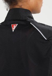 Czarna kurtka z teksturowanym siateczkowym tyłem, z czerwono-białym logo na kołnierzu i odblaskowymi akcentami na ramionach.