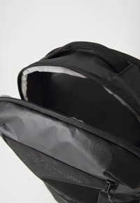 Svart ryggsekk med glidelåsåpning, teksturert overflate og indre rom. Har en logo på frontpanelet.