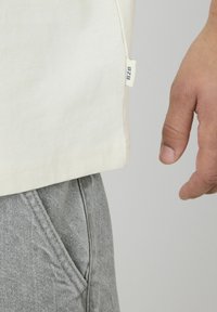 T-shirt crème clair à coupe droite, avec étiquette sur la couture latérale et texture subtile, associé à un pantalon en denim gris avec poches latérales.