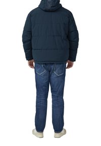Granatowa kurtka puchowa z kapturem, pikowana, z tłoczonym logo. W połączeniu z niebieskimi dżinsami i jasnymi trampkami, widziana od tyłu.
