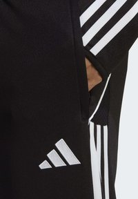 Black athletic pants with white three-stripe accents along the sides, featuring a side pocket and an embroidered logo near the hem.
