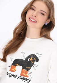 Sudadera crema con un diseño impreso de un dachshund negro con botas naranjas, con etiquetas que destacan características como "orejas caídas" y "ojos brillantes".