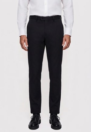Mężczyzna ubrany w czarne eleganckie spodnie, czarne skórzane buty i białą koszulę z długim rękawem, stojący na tle jednolitego tła.
