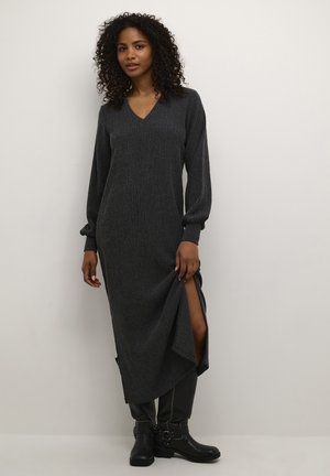 Robe tricotée longue gris foncé avec un décolleté en V et des manches longues, dotée d'échancrures latérales pour plus de liberté de mouvement. Associée à des bottes noires jusqu'aux genoux.