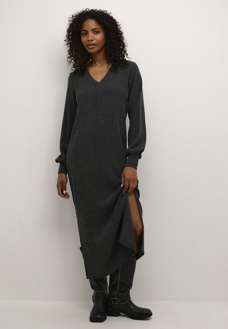 Robe tricotée longue gris foncé avec un décolleté en V et des manches longues, dotée d'échancrures latérales pour plus de liberté de mouvement. Associée à des bottes noires jusqu'aux genoux.