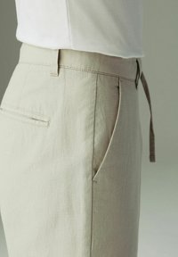 Pantalons de lin beige avec passants pour ceinture, poches à biais latérales et arrière, portés avec une chemise blanche rentrée sur un fond neutre.