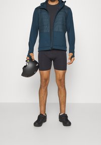 Veste matelassée bleue avec capuche, chemise noire ajustée, shorts noirs et casque noir tenu à la main. Le matériau semble respirant avec des accents texturés.