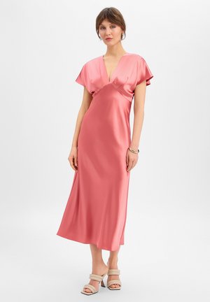 Vrouw die een mouwloze, zijden roze midi-jurk met V-hals draagt en beige sandalen met hakken, staand tegen een effen witte achtergrond.