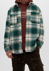 Giacca camicia a quadri verde e bianca con una texture pelosa, dotata di un taschino frontale e chiusura con bottoni, indossata sopra un felpa marrone.