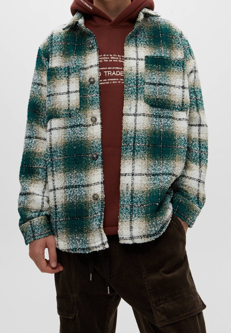 Giacca camicia a quadri verde e bianca con una texture pelosa, dotata di un taschino frontale e chiusura con bottoni, indossata sopra un felpa marrone.