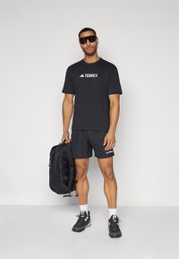 Sort atletisk t-shirt med "TERREX" logo, matchende sorte shorts, mørke sneakers og solbriller. Holder en sort rygsæk på den ene side.