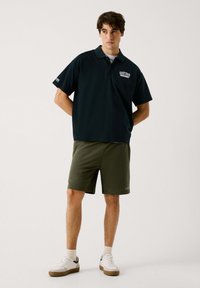 Donker marineblauwe poloshirt met korte mouwen en een kraag, voorzien van een wit logo. Gekoppeld aan olijfgroene shorts en witte sneakers.