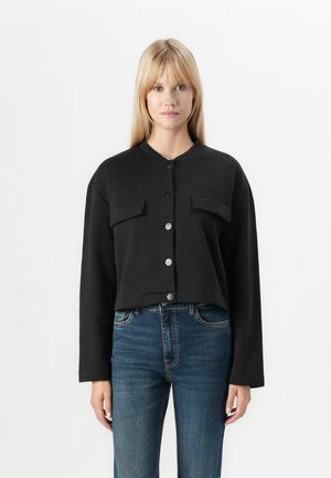 Black cropped jacket made of cotton, featuring a round neckline, long sleeves, two front pockets, and button closure. Worn with blue jeans.