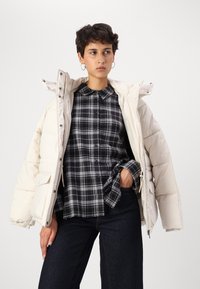 Padded cream jacket with hood, zip closure, and large front pockets, layered over a black and gray plaid button-up shirt and dark jeans.