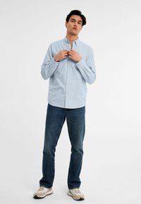 Lichtblauw verticaal gestreept overhemd met lange mouwen, gecombineerd met donkerblauwe jeans en beige sneakers. Eenvoudig, klassiek ontwerp.