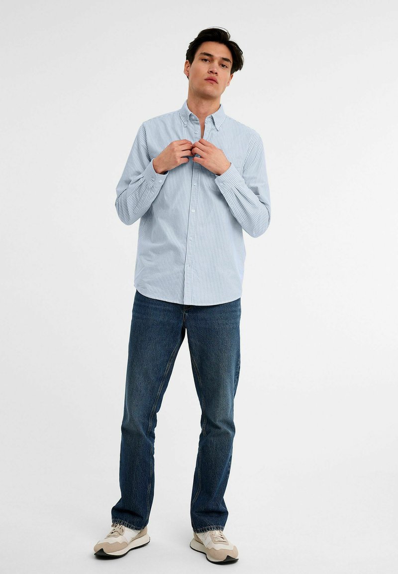 Lichtblauw verticaal gestreept overhemd met lange mouwen, gecombineerd met donkerblauwe jeans en beige sneakers. Eenvoudig, klassiek ontwerp.