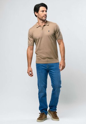 Mand iført beige kortærmet polo, blå jeans og brune sneakers, stående foran en ensartet lys baggrund.