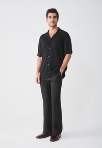 Camisa negra de manga corta con canalé vertical y botones, combinada con pantalones negros y zapatos burdeos. Diseño sencillo y moderno.