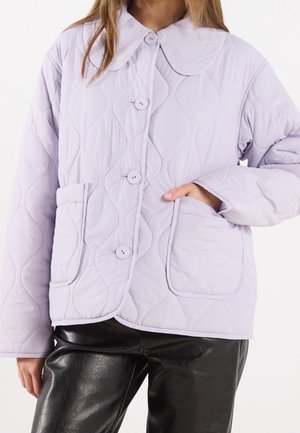 Femme portant une veste matelassée lavande avec de gros boutons, un col arrondi et deux poches plaquées à l'avant, associée à un pantalon en cuir noir.