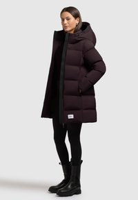 Abrigo tipo puff oscuro púrpura con cremallera negra y capucha. Presenta un diseño acolchado, bolsillos frontales y una etiqueta blanca en el dobladillo.