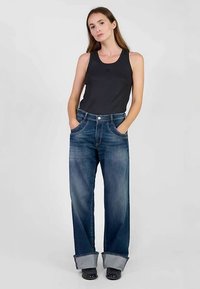 Camiseta sin mangas gris oscuro combinada con pantalones vaqueros de pierna ancha y color azul descolorido, con puños doblados y bragueta de botones. La modelo está de pie con las manos en los bolsillos.