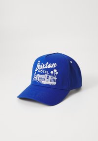 Blå basebollkeps med vit broderad text "THE Brixton MOTEL" och en grafik av en byggnad med palmträd på framsidan.