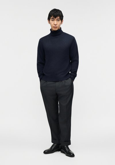 Μπλε ζιβάγκο πουλόβερ, με ριμπ μανστέτες, σε συνδυασμό με γκρι παντελόνι με λεπτές ρίγες. Το μοντέλο φοράει μαύρα δερμάτινα παπούτσια και στέκεται μπροστά σε απλό φόντο.