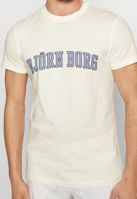 Kortärmad krämfärgad t-shirt i bomull med en iögonfallande blå "BJÖRN BORG"-grafik centrerad över bröstet. Slät textur.