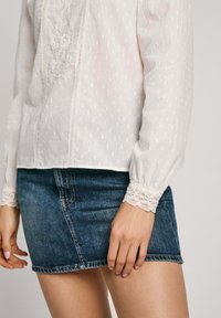 Blusa bianca ricamata con tessuto testurizzato, caratterizzata da pizzo ai polsini, abbinata a una mini gonna di jeans.